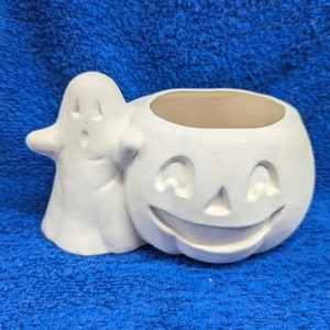 May include: White ceramic pumpkin with a ghost figure attached to the side. The pumpkin has a carved smiling face and a hollow top for holding items.