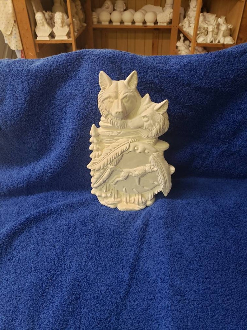 K&ouml;nnte beinhalten: Wei&szlig;e Keramikskulptur von zwei W&ouml;lfen mit einem Wolfswelpen, der vor ihnen l&auml;uft. Die W&ouml;lfe befinden sich in einer Waldkulisse mit B&auml;umen und Bl&auml;ttern.