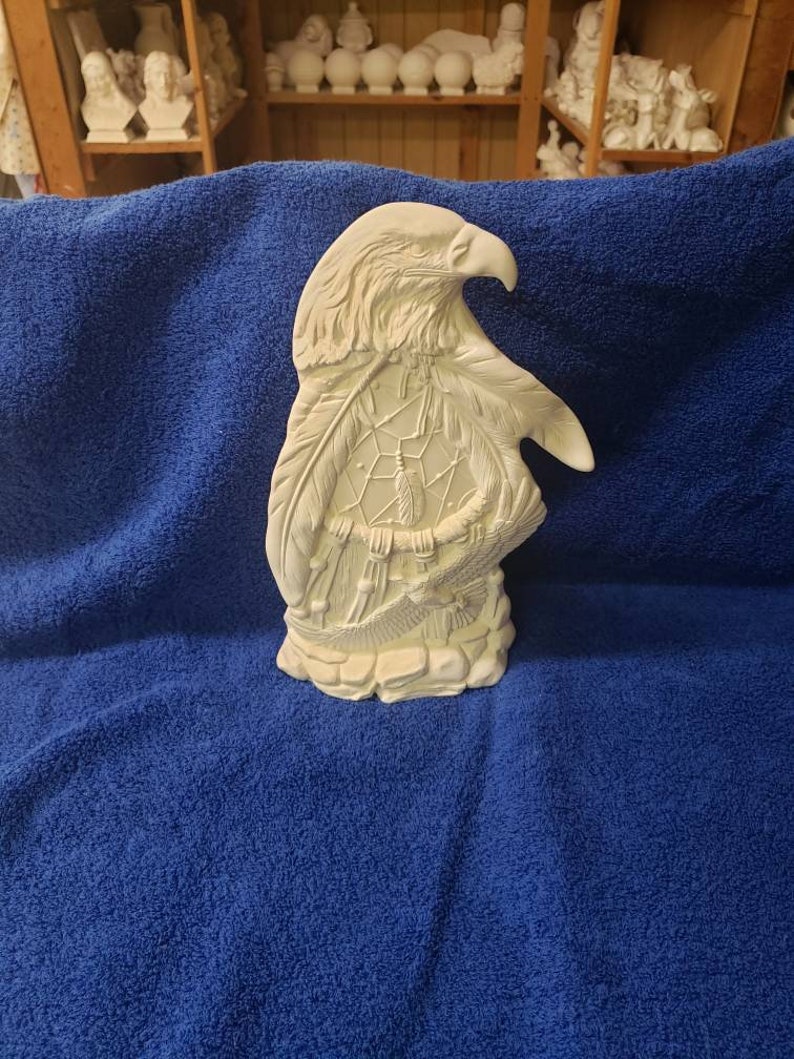K&ouml;nnte beinhalten: Wei&szlig;e Keramikfigur eines Adlers mit einem Traumf&auml;nger in seinen Krallen. Der Adler blickt nach rechts und hat seine Fl&uuml;gel ausgebreitet. Der Traumf&auml;nger besteht aus Federn und Perlen.