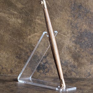 May include: A wooden pen with brass accents is displayed on a clear acrylic stand. The pen has a dark wood grain finish and a brass tip and top. The stand is angled, showcasing the pen against a brown background.