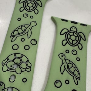 Può includere: Due cinturini per Apple Watch verdi con disegni di tartarughe e bolle nere. I cinturini sono in silicone e hanno una finitura liscia.