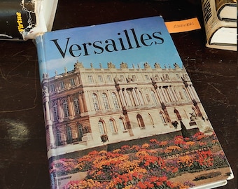 May include: A vintage hardcover book titled "Versailles" with a picture of the Palace of Versailles on the cover. The book is on a wooden table with a clock and other books.