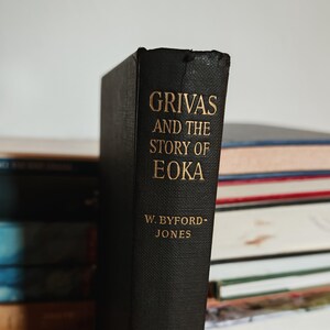 May include: A black hardcover book titled "Grivas and the Story of EOKA" with gold lettering. The author's name, W. Byford-Jones, is also in gold. The book is part of a stack of other books.