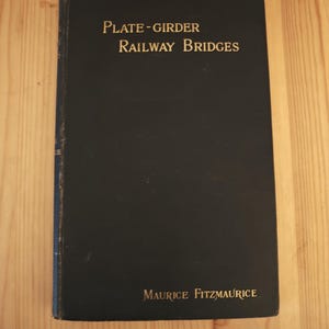 Può includere: Un libro d'epoca intitolato "Plate-Girder Railway Bridges" di Maurice Fitzmaurice. Il libro ha una copertina scura con scritte dorate. Il libro è appoggiato su una superficie di legno chiaro.