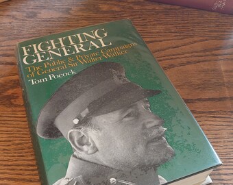 May include: A book titled "FIGHTING GENERAL" by Tom Pocock, with a portrait of General Sir Walter Walker on a green cover. Other books are stacked behind it. The book is on a wooden surface.