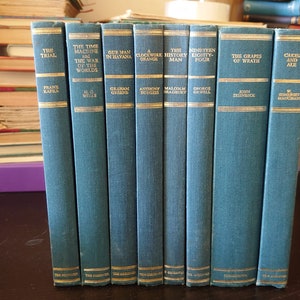 Könnte beinhalten: Eine Reihe von zehn blauen Hardcover-Büchern mit goldenen Schriftzügen auf den Rücken. Die Titel lauten "Der Prozess", "Die Zeitmaschine", "Unser Mann in Havanna", "A Clockwork Orange", "Der Geschichtsmann", "Neunzehn Achtzigvier", "Die Früchte des Zorns", "Kains und Abel".