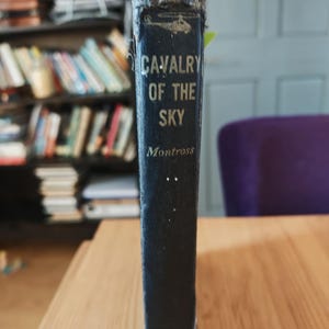 May include: A vintage book titled "Cavalry of the Sky" by Montross. The book has a dark blue cover with gold lettering and a small illustration of a helicopter. The spine shows signs of wear and tear, with frayed edges. The book is on a wooden surface.