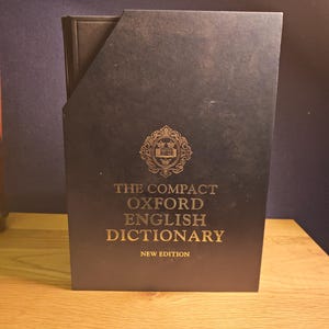Peut inclure: Un livre marron foncé avec des lettres dorées et un emblème décoratif. Le texte sur la couverture indique "THE COMPACT OXFORD ENGLISH DICTIONARY NEW EDITION". Le livre est dans un étui assorti.