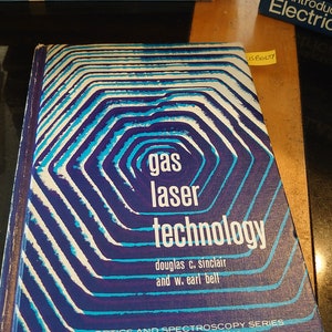May include: A hardcover book titled "gas laser technology" with a blue and white geometric design on the cover. The book is part of the Optics and Spectroscopy Series. Other books are stacked in the background.