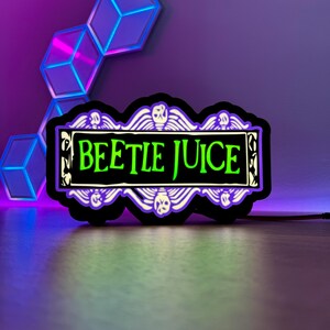 May include: A black and white neon sign with the words "Beetle Juice" in green letters. The sign has a decorative border with wings and skulls.