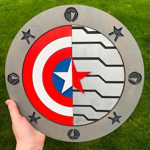 Puede incluir: Un letrero redondo de madera con un diseño de escudo de Capitán América en una mitad y un logotipo de Capitán América en la otra. El escudo es rojo, blanco y azul con una estrella en el centro. El logotipo es una estrella blanca sobre un fondo azul. El letrero está rodeado por un círculo de ocho estrellas.