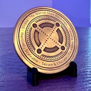 May include: A gold-colored metal coin with the text "Elrond - The Internet Scale Blockchain - Massively Scaleable - Secure & Efficient - Developer Friendly" around the edge. The center of the coin features a stylized design with four circles connected by lines.