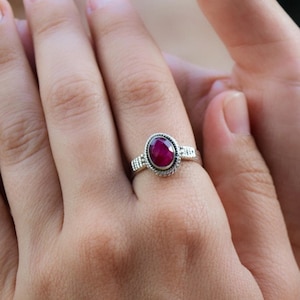 Könnte beinhalten: Ein silberner Ring mit einem tief roten ovalen Edelstein, der in einer Zargenfassung gefasst ist. Der Ring hat eine strukturierte Band.