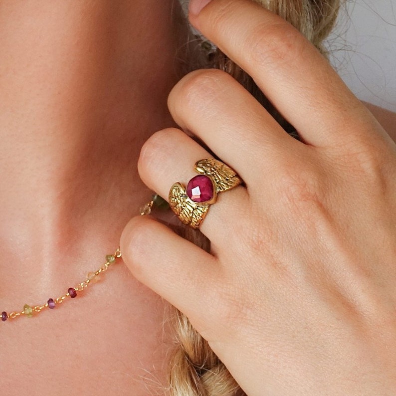 Puede incluir: Anillo de oro con una piedra preciosa roja engastada en el centro, rodeada de dise&ntilde;os de alas detallados.