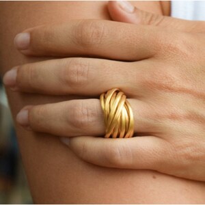 Puede incluir: Un anillo dorado con un diseño tejido. El anillo se lleva en un dedo.