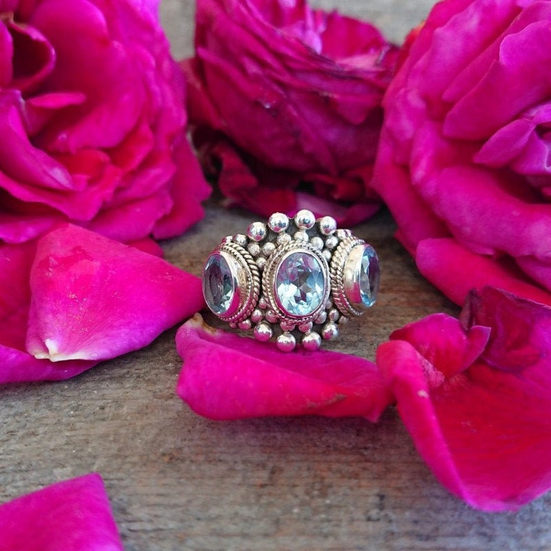 May include: A silver ring with three blue topaz gemstones set in a beaded band. The ring is surrounded by pink rose petals.