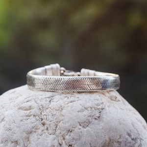 Peut inclure: Un bracelet en argent avec un motif en chevrons. Le bracelet est posé sur une pierre blanche.