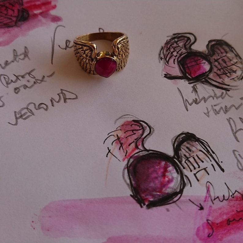 Puede incluir: Un anillo de oro con una piedra preciosa roja rub&iacute; y dos alas a cada lado de la piedra.