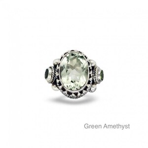 May include: A silver ring with a large oval green amethyst gemstone surrounded by smaller green gemstones. The ring has a detailed silver band.