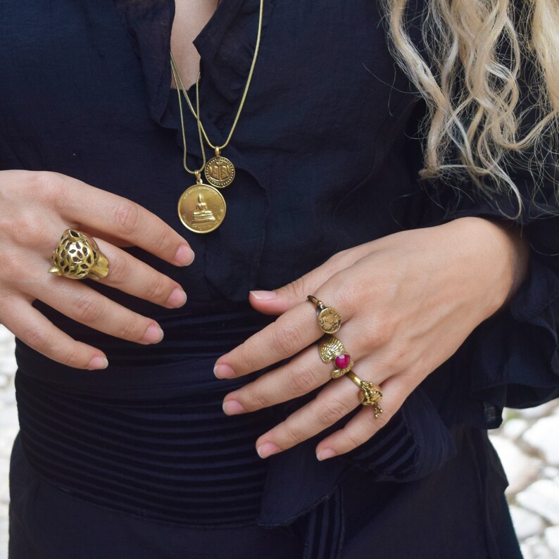 Puede incluir: Una mujer lleva una blusa negra con un collar de cadena dorada con tres colgantes de monedas de oro. Lleva un anillo de oro con un dise&ntilde;o de cabeza de leopardo en su mano izquierda y tres anillos de oro en su mano derecha, uno con una piedra roja.