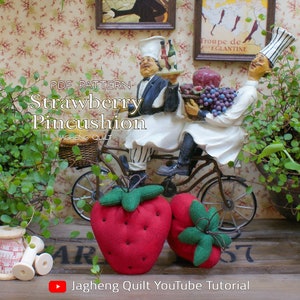 May include: Two red strawberry pincushions with green fabric leaves and black seed details. The text "Strawberry Pincushion" is displayed. A decorative figurine of two chefs on a bicycle is in the background. The text "Jagheng Quilt YouTube Tutorial" is also visible.