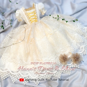 May include: A white lace doll dress with yellow ribbon detailing and a ruffled skirt. The dress is on a white fabric background with green vines. The text "PDF PATTERN Hana's Dress & Shoes" is on the dress. The text "Jagcheng Quilt YouTube Tutorial" is below the dress.