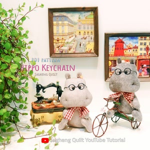 May include: PDF pattern for a grey fabric hippo keychain with glasses and a red and white checked scarf. The hippo is sitting on a table with a sewing machine and a small wooden box. There are two paintings in the background, one of a city street and one of a windmill.