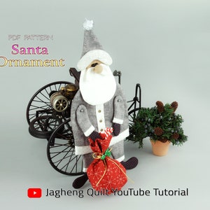 Puede incluir: Adorno de Papá Noel hecho a mano, con abrigo y gorro grises, barba blanca. Sostiene una bolsa de regalo roja con un lazo dorado. Se muestra el texto "Santa Ornament". Un coche antiguo en miniatura y una pequeña planta en maceta están en el fondo.
