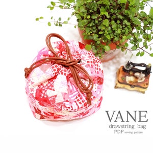 K&ouml;nnte beinhalten: Ein runder Kordelzugbeutel aus Patchwork in Rot- und Rosat&ouml;nen, mit braunem Kordelverschluss. Der Beutel hat eine gesteppte Optik. Der Text auf dem Bild lautet "VANE Kordelzugbeutel PDF Schnittmuster".