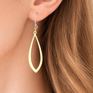 May include: A gold-colored, open teardrop earring is shown. The earring hangs from a clear plastic hook. The earring is a simple, elegant design, with a smooth, polished surface. The background is blurred, showing a person's ear and hair.