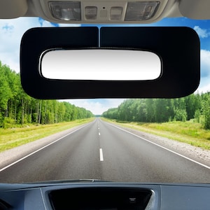 May include: A black car sunshade with a built-in mirror. The sunshade is attached to the rearview mirror of the car. The sunshade is blocking the sun from shining into the car. The sunshade is in a car with a view of a road through a forest.