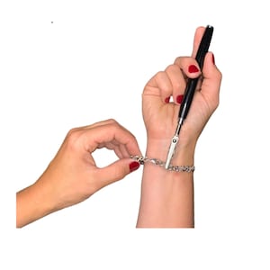 May include: A silver bracelet with a lobster clasp is being fastened with a black pen-like jewelry tool. The tool has a clip at the end to hold the bracelet. The person's nails are painted red.