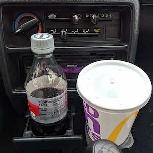 May include: The interior of a black car with a clear bottle of soda in a cup holder, a white cup with a purple and yellow logo, and a black gear stick.