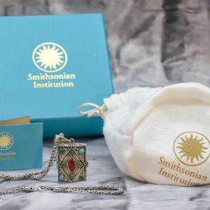 May include: A silver necklace with a small, ornate box pendant. The box is decorated with colorful stones and has a sun design on the front. The necklace comes in a blue box with the Smithsonian Institution logo and a white pouch with the Smithsonian Institution logo.