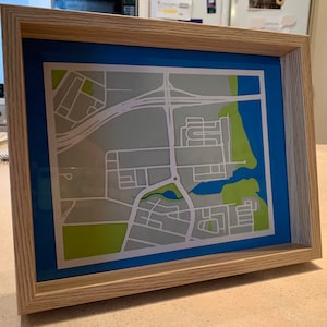 May include: A framed map of a neighborhood with a blue mat and a light wood frame. The map is printed in shades of gray and green, with a small lake in the center.