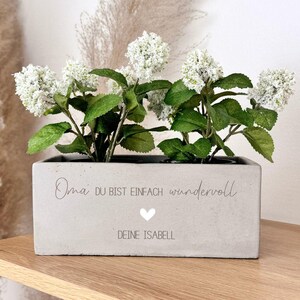 Könnte beinhalten: Ein rechteckiger Beton-Pflanzkübel mit weißen Blüten und grünen Blättern. Der Pflanzkübel trägt den Text "Oma du bist einfach wundervoll" und "DEINE ISABELL" mit einem Herzsymbol. Der Pflanzkübel steht auf einem Holzregal.