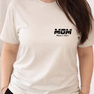 Könnte beinhalten: Cremefarbenes T-Shirt mit dem Wort "MOM" in fetten, schwarzen Buchstaben und den Namen "Melia & Anton" darunter. Das Shirt hat kurze Ärmel und eine lockere Passform.