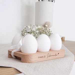 Op de afbeelding: Houten eierhouder met een witte basis en een natuurlijke houten bovenkant. De houder heeft vier vakken voor eieren en is gegraveerd met de tekst "Familie Schneider".