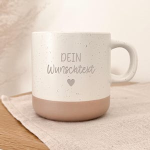 Può includere: Una tazza in ceramica bianca con finitura maculata e base marrone chiaro. La tazza presenta il testo "DEIN Wunschtext" e un piccolo cuore. La tazza è su un tovagliolo di lino beige.