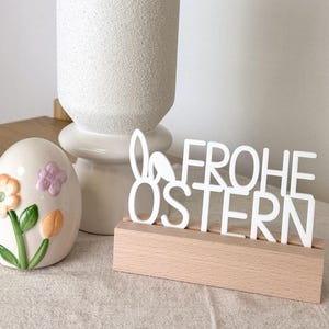 Könnte beinhalten: Eine weiße Keramikvase mit Kätzchenzweigen, ein dekoratives Ei mit floralen Designs und ein Holzschild mit der Aufschrift "FROHE OSTERN" in weißen Buchstaben. Das Schild hat Hasenohren in das Design integriert.