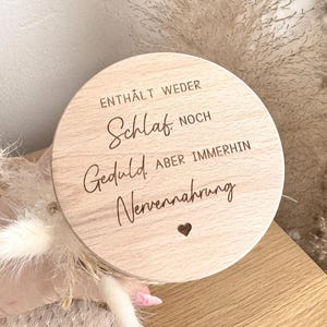May include: A round wooden sign with engraved text in German. The text reads "ENTH&Auml;LT WEDER Schlaf NOCH Geduld ABER IMMERHIN Nervennahrung" with a small heart symbol. The sign is light brown and held in a hand, with dried flowers in the background.