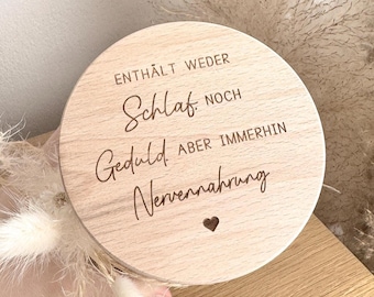 May include: A round wooden sign with engraved text in German. The text reads "ENTHÄLT WEDER Schlaf NOCH Geduld ABER IMMERHIN Nervennahrung" with a small heart symbol. The sign is light brown and held in a hand, with dried flowers in the background.