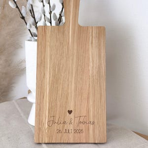 Könnte beinhalten: Rechteckiges Schneidebrett aus hellem Holz mit Griff und Aufhängeloch. Auf dem Brett sind die Namen "Julia & Tobias", ein Herzsymbol und das Datum "26. JULI 2025" eingraviert.
