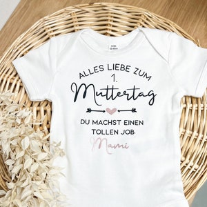 Könnte beinhalten: Weißer Baby-Body mit kurzen Ärmeln. Der Body hat den Text "ALLES LIEBE ZUM 1. MUTTERTAG" in schwarzer Schrift, mit "DU MACHST EINEN TOLLEN JOB Mami" in schwarzer und rosa Schrift. Der Body liegt auf einem geflochtenen Korb.