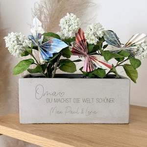 Op de afbeelding: Een rechthoekige betonnen plantenbak met witte bloemen en origami vlinders in blauw, rood en groen. De plantenbak heeft de tekst "Oma, DU MACHST DIE WELT SCHÖNER, Max Paul & Lena."