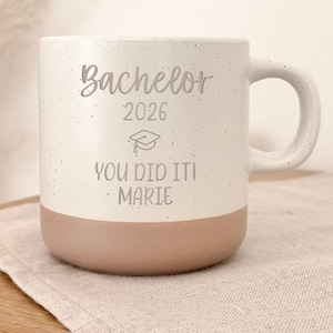 Op de afbeelding: Een witte keramische mok met een gespikkelde afwerking en een lichtbruine basis. De mok heeft een handvat en heeft de tekst "Bachelor 2026" met een afstudeerpet-afbeelding en de woorden "YOU DID IT! MARIE".