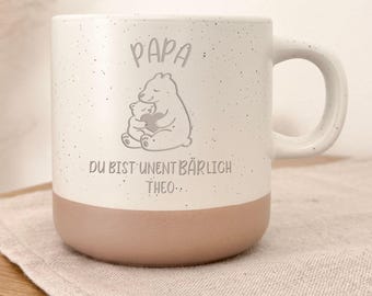 May include: A white ceramic mug with a speckled finish and a light brown base. The mug features a gray illustration of a bear hugging a smaller bear, with the German text "Papa, Du bist unentbärlich Theo..." printed above and below the illustration.