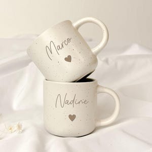 Könnte beinhalten: Zwei cremefarbene Keramiktassen mit gesprenkelter Oberfläche. Die obere Tasse zeigt den Namen Marco in Schreibschrift über einem kleinen Herz. Die untere Tasse zeigt den Namen Nadine in Schreibschrift über einem kleinen Herz. Beide haben einen Henkel.