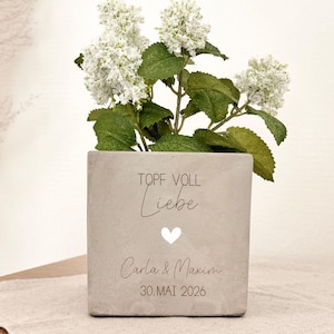 Pode incluir: Um vaso quadrado de concreto cinza claro com flores brancas e folhas verdes. O vaso tem as palavras "TOPF VOLL Liebe" e os nomes "Carla & Maxim" com a data "30.MAI 2026" impressos. Um coração branco também é impresso.