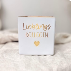Può includere: Un portacandele quadrato bianco con scritta dorata "Lieblings KOLLEGIN" e un cuore dorato. Il bordo superiore del portacandele è dorato. Un lumino è incluso.